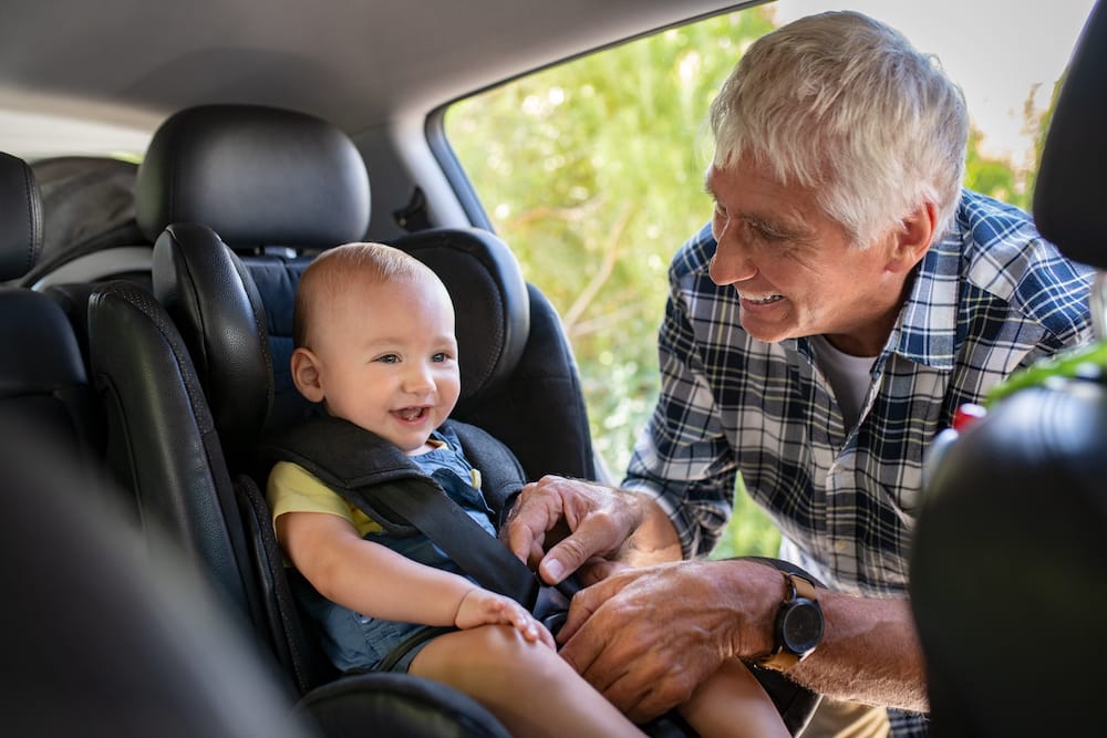 Grandfather tying baby in child seat. Cute little boy going for road trip with grandfather. Happy senior man and happpy smiling grandchild enjoying car trip. Toddler boy buckled into car seat.