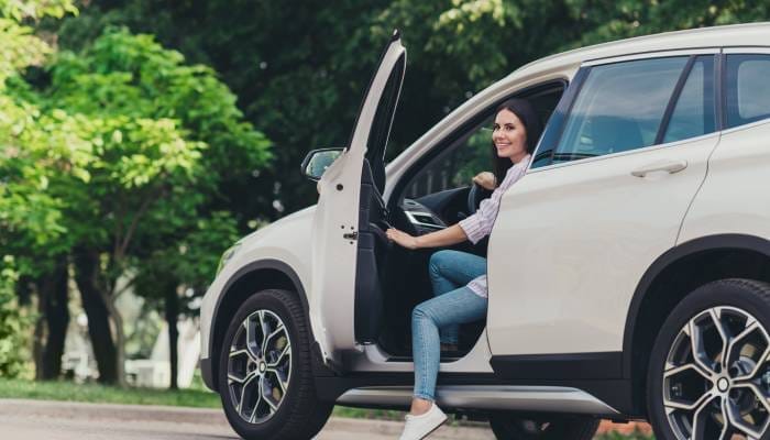 A young woman opening the driver-side door and stepping out of her white midsize van in a parking spot for a park.