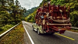 An overloaded pickup truck holds a large stack of wooden furniture tied down with strands of thin rope.