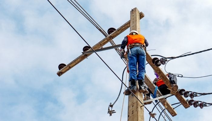 Two electrical workers on top of a wooden electrical pole. They are wearing protective hats and other protective gear.