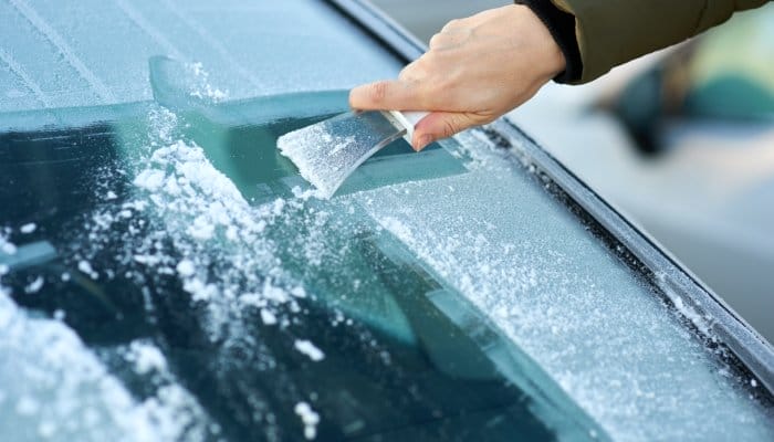 A close-up of someone using an ice scraper to remove ice from a car windshield covered in snow and ice.