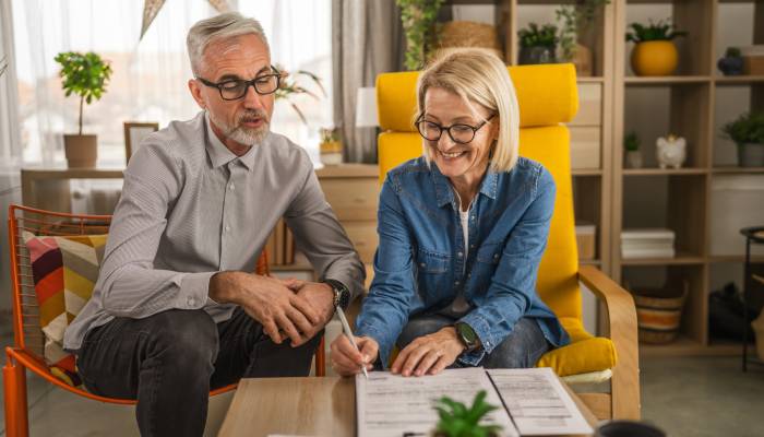 A mature man and woman sit in a cozy room, reviewing documents. The woman smiles as she prepares to sign a document.