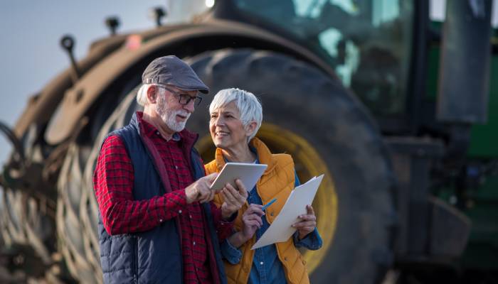 An older couple wearing winter vests stand in front of a large tractor. The man looks at a tablet and the woman holds papers.