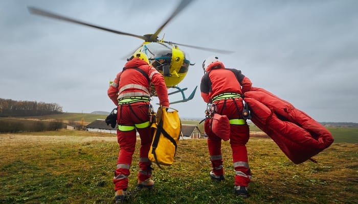 Two paramedics wearing red jumpsuits and helmets making their way toward a helicopter to perform a rescue operation.