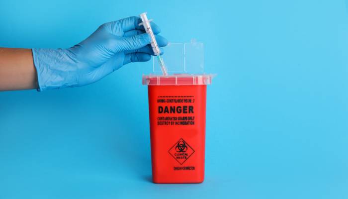 A blue-gloved hand is dropping a used syringe into a bright red sharps container for later disposal.
