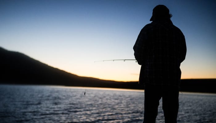 A person holding a rod and reel is fishing at sunset on a lake with mountains or hills ahead in the distance.
