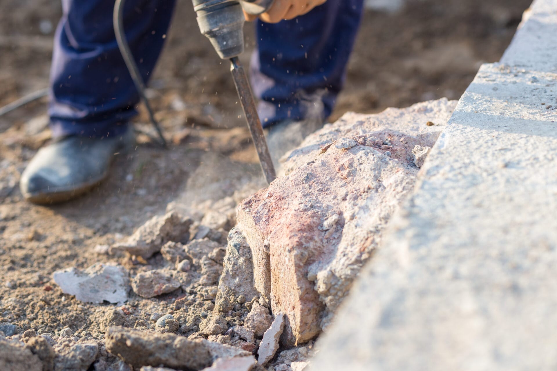 A close-up of a worker holding a jackhammer to destroy a mound of concrete next to a wall on a construction site.