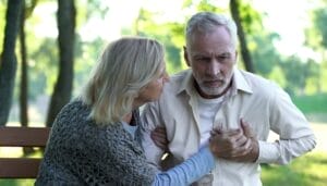 Home 16 An older adult couple sitting on a bench outside. The man is clutching his chest and the woman appears concerned.