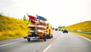 A mustard-yellow van is towing eight kayaks on the back of the vehicle. It's driving along a busy highway.