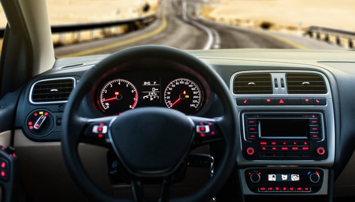 A vehicle's dashboard is illuminated with red and white lights. The vehicle is driving down a road surrounded by desert.