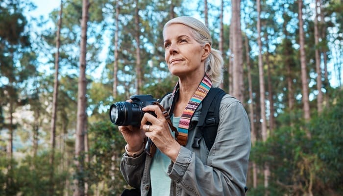 A woman with white hair smiles and holds a DSLR camera as she stares off into the distance in a forest.