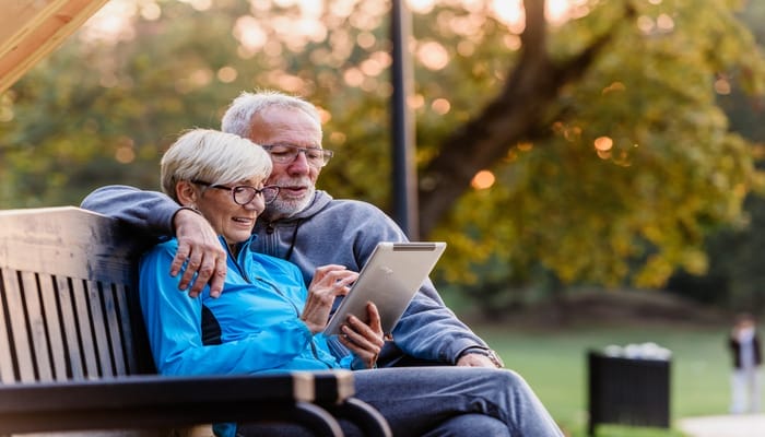 A boomer couple sitting on a bench outside enjoying the fresh air. They're staring and smiling at a mobile device.