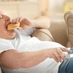 Home 5 A bald, elderly man lying on a couch while eating a hamburger. He also holds a remote in his other hand.