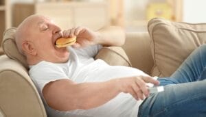 A bald, elderly man lying on a couch while eating a hamburger. He also holds a remote in his other hand.