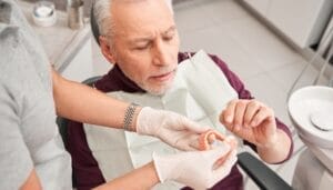 A senior man sitting in a dentist chair while an individual in white gloves shows him a pair of dentures.
