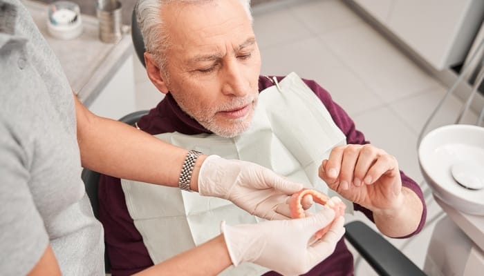 A senior man sitting in a dentist chair while an individual in white gloves shows him a pair of dentures.
