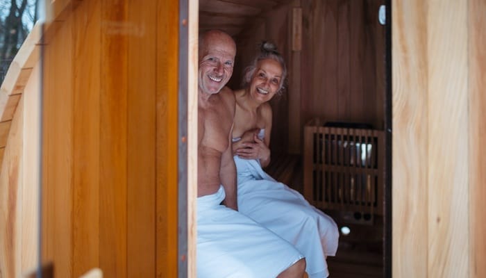 A happy senior couple sit on a bench in a private wooden sauna with a glass door. They both wear white towels.