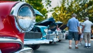 Several cars are lined up. The foremost one has red paint and circular headlights. There is a group of men in the background.