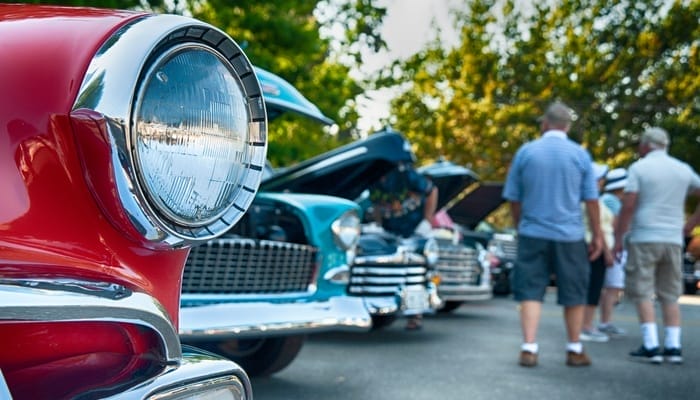 Several cars are lined up. The foremost one has red paint and circular headlights. There is a group of men in the background.