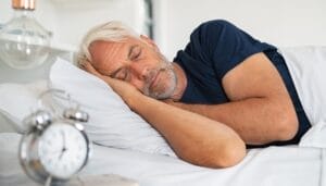 A man with white hair in a blue shirt sleeping on his side in a bed with white bedding with an alarm clock in the foreground.