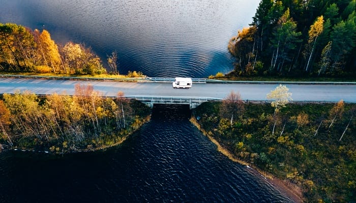 An aerial view shows a white RV crossing a short bridge. The bridge crosses a body of water that's surrounded by fall foliage.