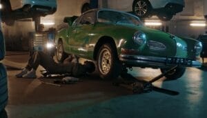 A mechanic working on the undercarriage of a green vintage car lifted on a jack in an automotive workshop.