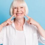 Home 2 A woman with a short white bob and a white shirt grins as she points to her smile, showing off her dentures.