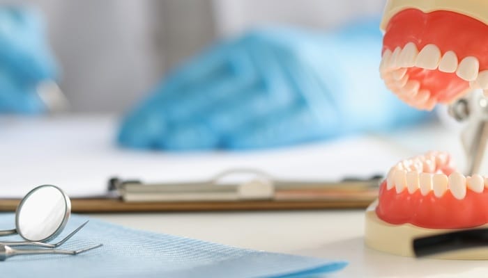 Home 1 A close-up of a full denture set sits on a desk with dental tools. A dentist sits in the background with blue gloves on.
