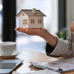 Home 11 Someone holds a small wooden model of a home in one hand and keys in the other. A laptop, a calculator and a clipboard are on a desk.