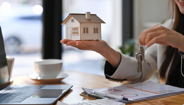 Someone holds a small wooden model of a home in one hand and keys in the other. A laptop, a calculator and a clipboard are on a desk.