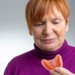 A woman with a pixie cut wearing a purple turtleneck, looking down at a set of dentures in her hand.
