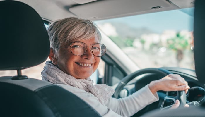 An older woman sitting in the driver seat of a car. She is looking in the backseat with one hand on the wheel.