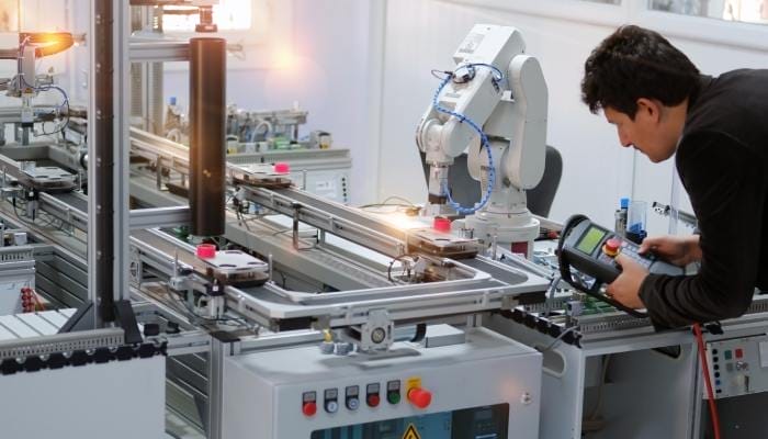 An employee in a factory is holding a handheld device in front of an automated robotic arm attached to a machine.