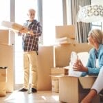 A smiling couple packing boxes in a bright, modern home with large windows, wooden floors, and scattered moving boxes.
