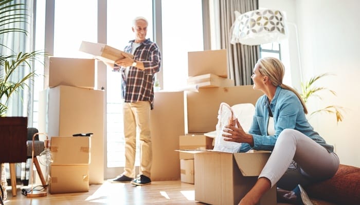 A smiling couple packing boxes in a bright, modern home with large windows, wooden floors, and scattered moving boxes.