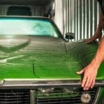 A man with rolled-up sleeves is standing next to a green car that appears to be in a room with metal walls.