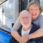 A senior couple laughing and playfully embracing in front of their RV that is parked at a forest campground.