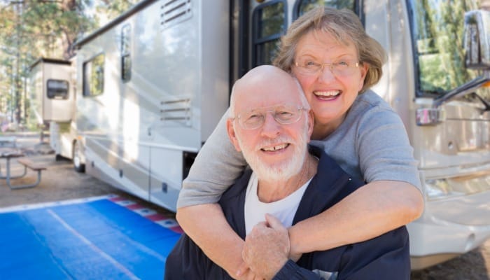 A senior couple laughing and playfully embracing in front of their RV that is parked at a forest campground.