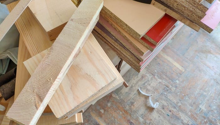 Pile of wood offcuts and boards on a workbench, with plywood and particleboard sheets over a worn floor.